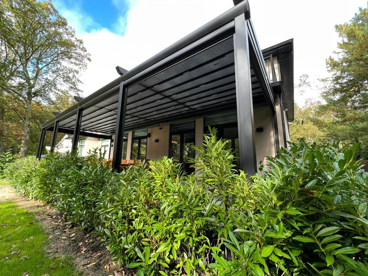 A dark retractable fabric-roof structure beside a house, viewed through garden planting.