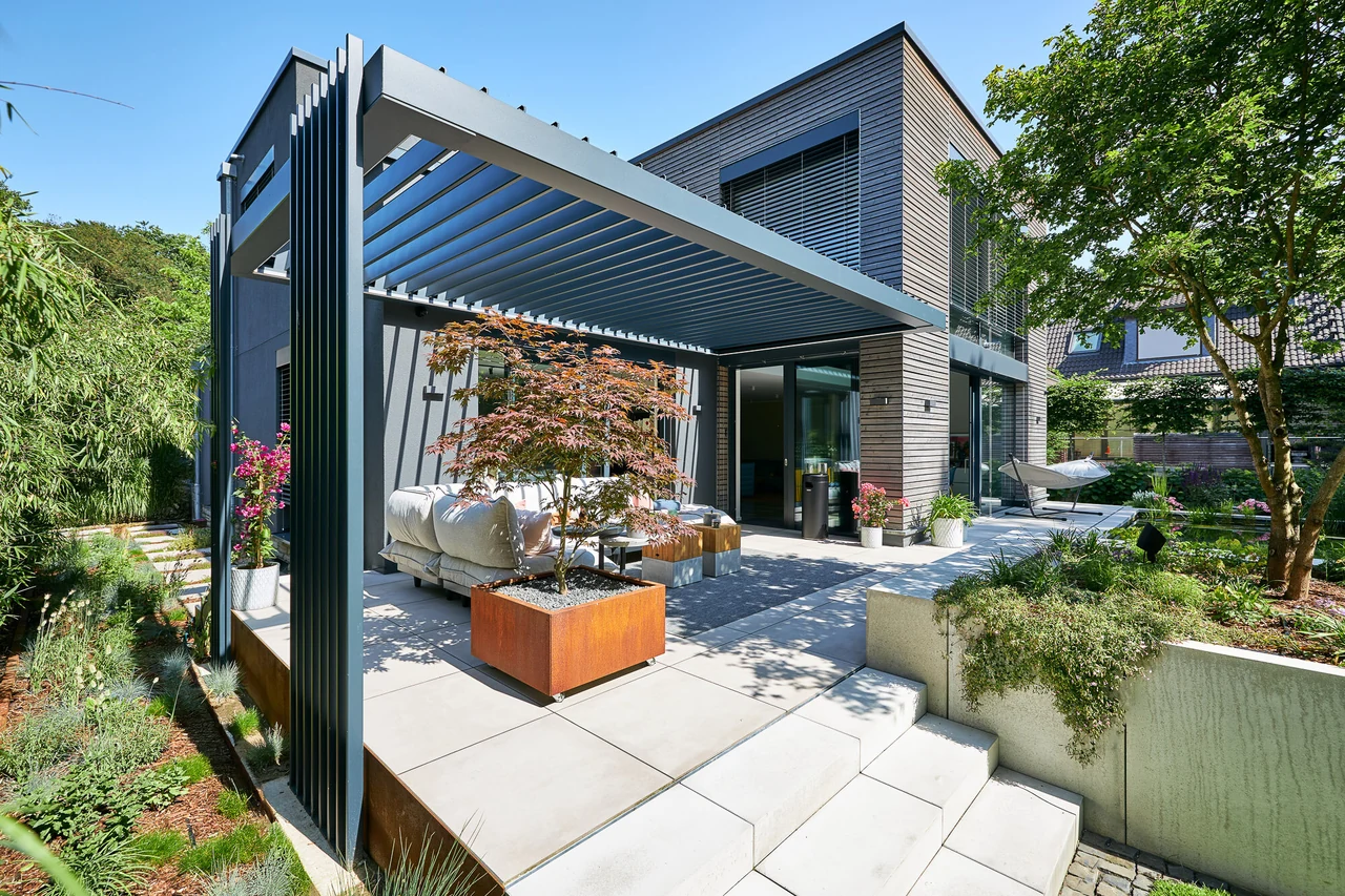 Dark louvered pergola beside a two-storey house, with planted borders and garden seating.